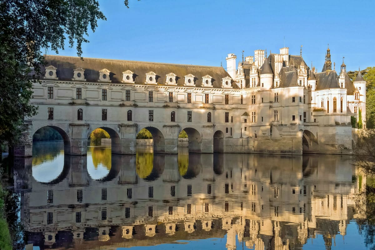 The Château de Chenonceau spanning the River Cher — five white stone arches across the water, steep slate roof with Renaissance turrets, reflected in the still river. Loire Valley, France.